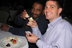 Two men sitting at a table eating desserts.