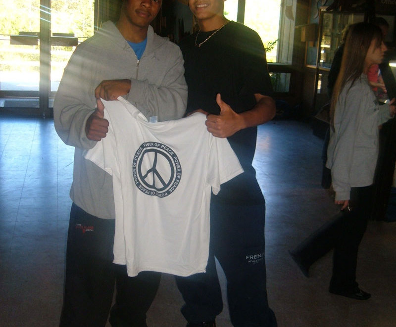 Two young men standing next to each other holding a peace sign t - shirt.